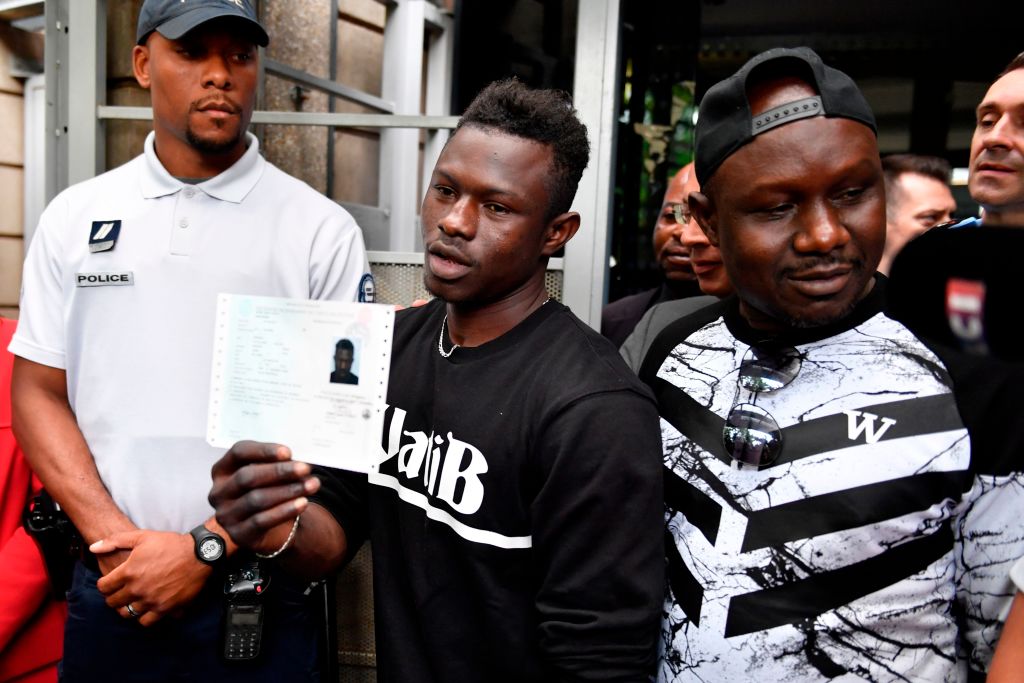 Malian migrant Mamoudou Gassama (C) flanked by his older brother (R) holds his temporary residence permit after receiving it at the Prefecture of Bobigny, northeast of Paris on May 29, 2018, one day after he was honored by the French President for scaling an apartment building to save a 4-year-old child dangling from a fourth-floor balcony. - Two days after his daring rescue -- viewed millions of times online -- Mamoudou Gassama, 22-year old, nicknamed "Spiderman" by French media for his astonishing climbing ability and feted as a hero, was offered by French President citizenship, and a job with the fire service.