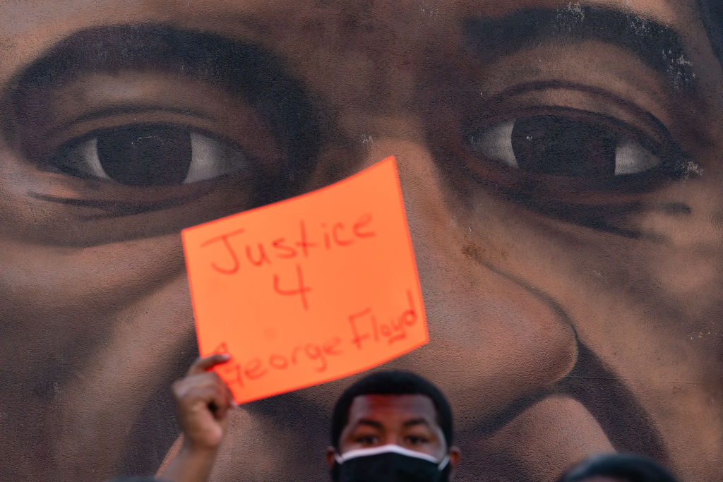 A man holds up a sign in front of a mural of George Floyd f