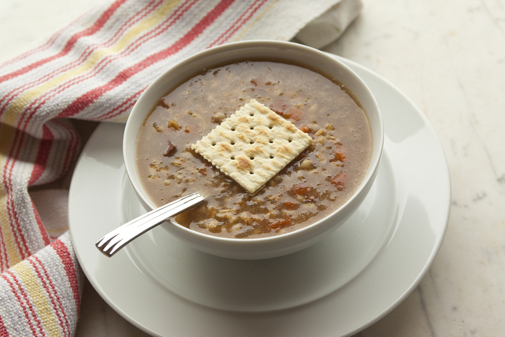 Bowl of vegetarian barley soup with a salt cracker garnish in a white bowl with saucer on a white marble table