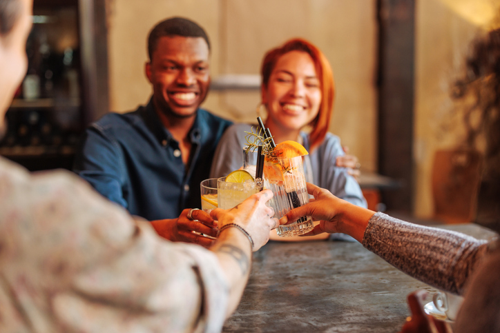 Young African-American man and Hispanic woman toasting cocktail glasses with friends in restaurant.