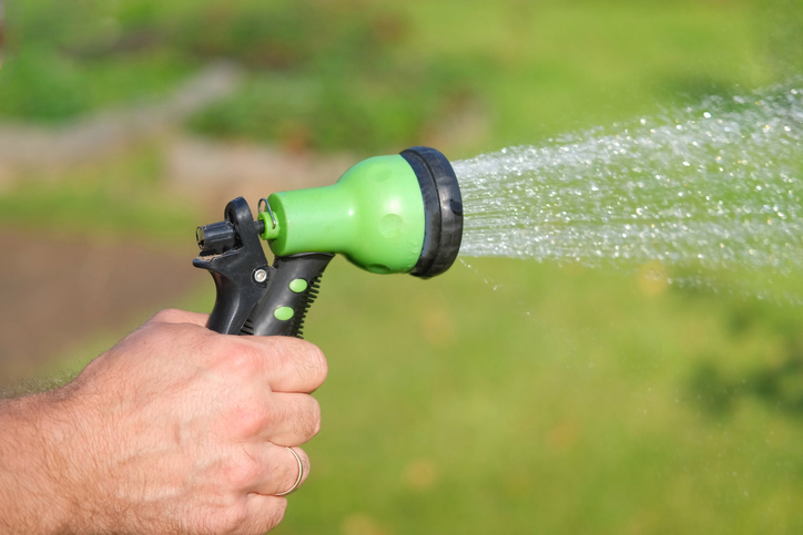 Gardener with a watering hose. Person spraying green grass lawn with hose sprayer. Irrigation with water, sunny day. Garden sprinkler in action. Landscaping. Gardening, waters, growing and plants care