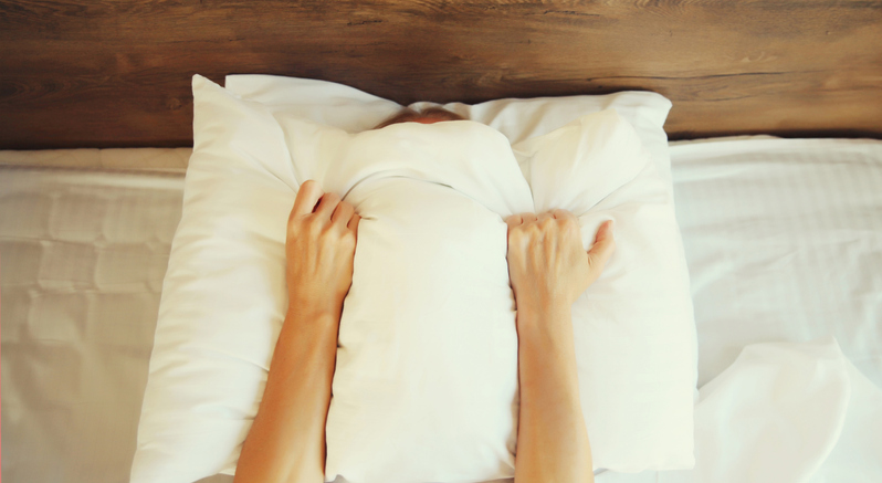 Unhealthy sleep, woman suffering from insomnia lying on the bed and holding in hands covering her head with white pillow in bedroom at home