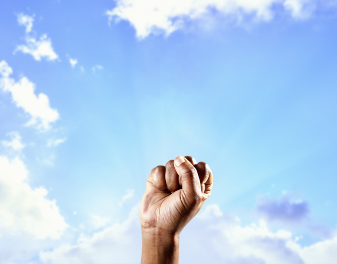 Man's clenched fist raised against a summer sky with sun's rays emerging from behind a cloud