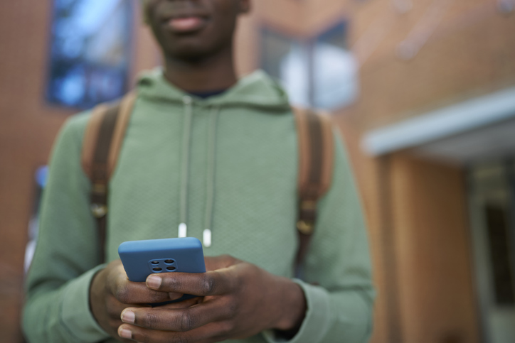 Close Up Of Male Teenage High School College Or University Student Checking Social Media Or Text Messaging On Mobile Phone Outside Building